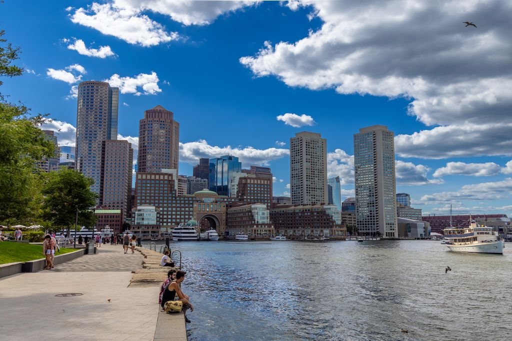 Explore the vibrant Boston harbor front with towering skyscrapers and clear blue skies reflecting on the water.